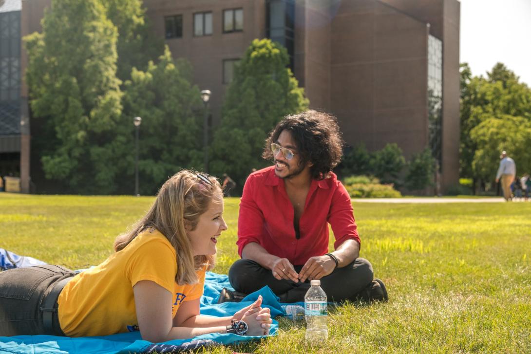 A male and a female student relax outdoors on the grass in front of a building on NEIU's Main Campus