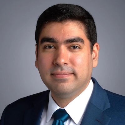 A headshot photo of Amirhossein Maleki, smiling, wearing a dark blue suit, white collared shirt and blue tie, against a gray background.