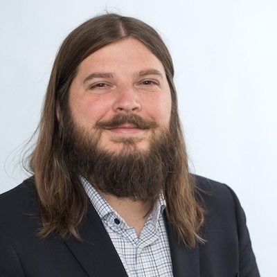 A headshot photo of Andrew Young smiling, wearing a white collared shirt with a colorful plaid pattern and black blazer against a white/light gray background.