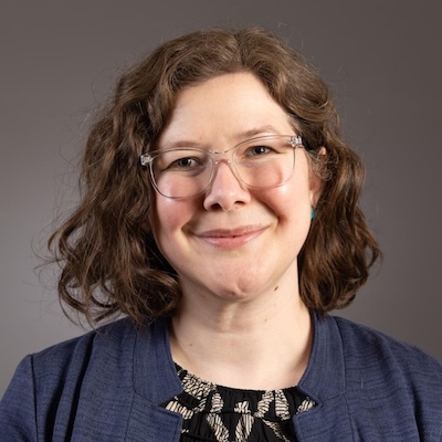 A headshot photo of Ashley L. Elrod smiling, wearing clear rimmed glasses, a black printed top and blazer against a gray background.