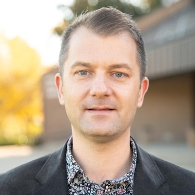 A headshot photo of Christopher Straughn, wearing a floral button down shirt and black blazer. The Ronald Williams Library is blurred in the background.