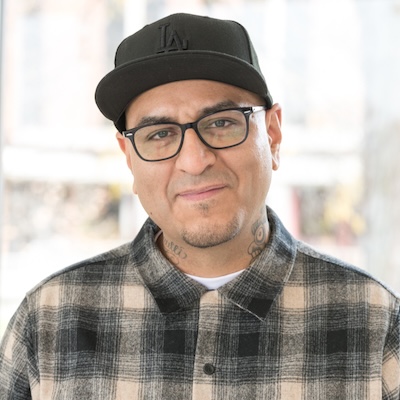 A headshot photo of J. Adrian Castrejon smiling, wearing a black LA Dodgers baseball hat, black rimmed glasses and tan and gray plaid flannel button down shirt.