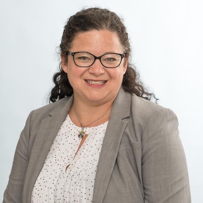 A headshot photo of Jody Siker, smiling, wearing a white top with polka dots, gray blazer and black rimmed glasses.
