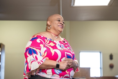 A photo of Gwendolyn Baxter, smiling, wearing a pink blouse with a floral pattern in the library at Northeastern Illinois University's Jacob H. Carruthers Center for Inner City Studies.