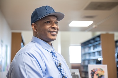 A photo of LeVon Stone Sr. wearing a Chicago Bears flat brim cap, light blue shirt and blue tie, smiling in the library at Northeastern Illinois University's Jacob H. Carruthers Center for Inner City Studies.