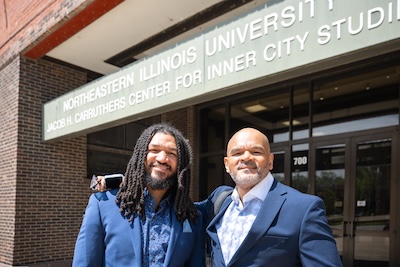 A photo of Jerome Anderson II (left) and Jerome Anderson Sr. (right) in front of Northeastern Illinois University's Jacob H. Carruthers Center for Inner City Studies, smiling, wearing blue suits on a sunny day.