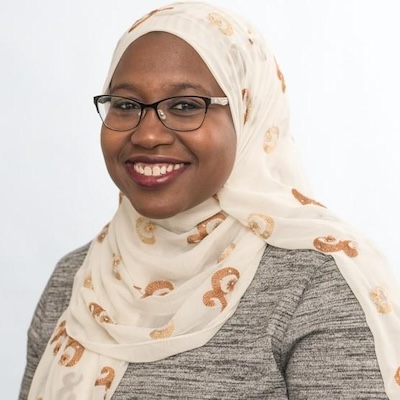 A headshot photo of Olunbunmi B. Oyewuwo-Gassikia, smiling, wearing brown and tan rimmed glasses, a gray top and white hijab with a gold pattern.