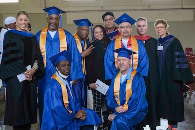 Five graduates wearing blue and gold regalia stand surrounded their mentors and NEIU admin and faculty
