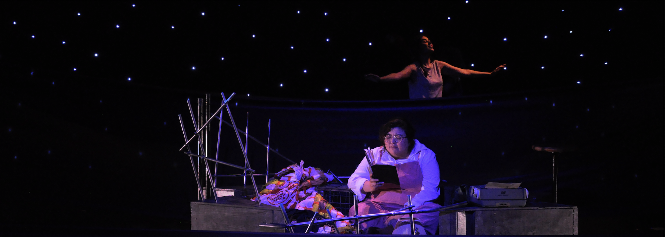 in the belly of the whale: an actor sits on a boat writing and behind them is another actor swimming in the open ocean