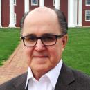 Dr. Frederick Prete is wearing black glasses and a white collared shirt as he stares at the camera in front of a red building and a grassy lawn