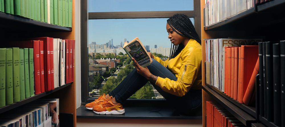 A female student reads a book while seated on the interior ledge of a window on the Carruther Center building.