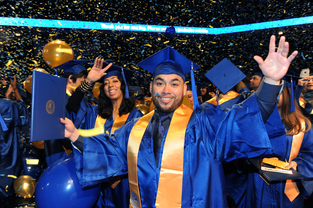 A smiling male graduate wearing a blue robe holds up his diploma at the December 2016 Commencement ceremony