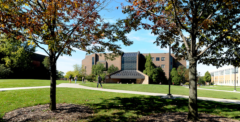 Students walk the paths of the University Commons with the Library in the background and two trees in the foreground