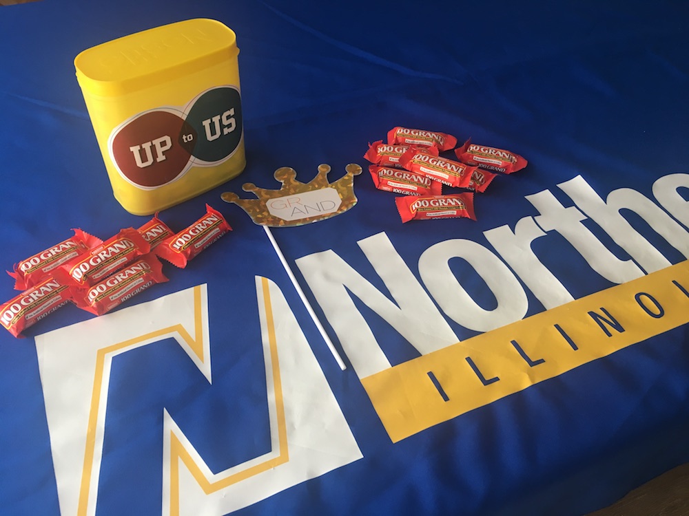 An Up to US branded yellow plastic box rests on an NEIU branded tablecloth amid a scattering of 100 Grand chocolate bars