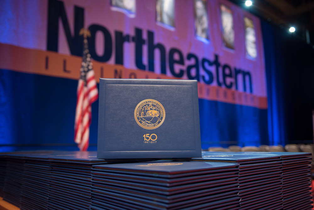 A diploma cover on display at December 2017 Commencement with an American flag and a branded Northeastern banner in the background
