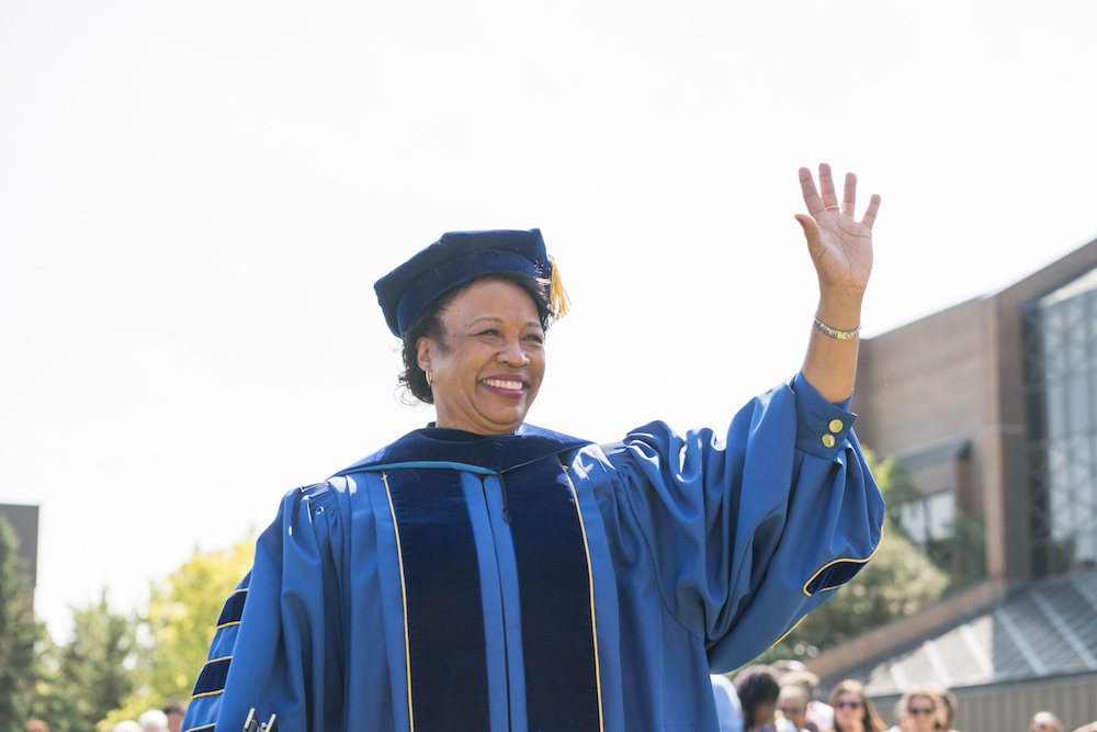 President Gloria Gibson, wearing academic regalia, raising her hand in greeting
