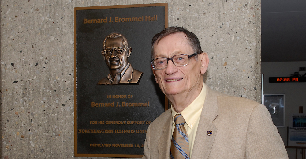 Bernard J. Brommel stands in front of the plaque in Bernard Brommel Hall that bears his name and likeness