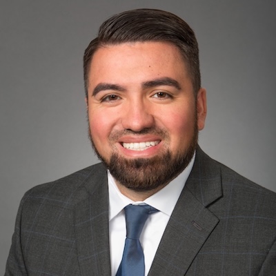 A headshot photo of Jose "Jay" Fulgencio smiling, wearing a gray suit, white collared shirt and blue tie against a gray background.
