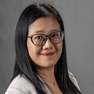 A headshot photo of Xueijing "Jessie" Bai, smiling, wearing brown glasses and a light gray blazer against a dark gray background.