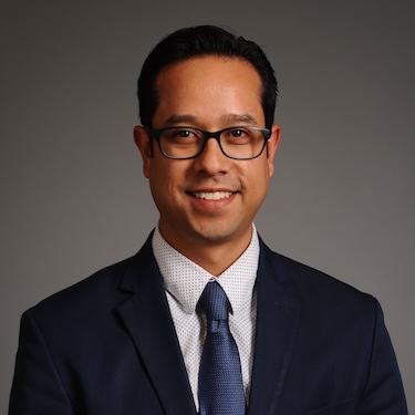 A headshot photo of Aaron Cortes wearing dark colored suit with blue tie and collard shirt and glasses, smiling against a gray background.