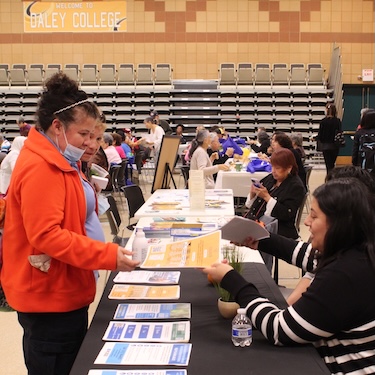 A person in an orange coat is handed a flyer from a person in a black and white striped sweater sitting behind a table during the ChicagoCHEC Community Forum in 2023. 