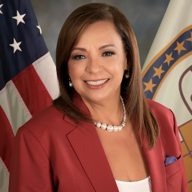 A headshot photo of Iris Y. Martinez, smiling, wearing a red blazer with white blouse and white necklace. The flags of the United States and Cook County are visible in the background.