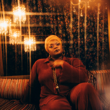 A photo of Kyshona wearing a maroon outfit, sitting on a striped couch, against a floral background. The photo is accented in warm lighting tones. A few chandeliers are also visible in the background. Photo by: Anna Haas