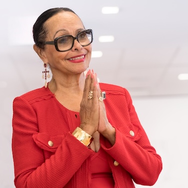 A photo of Murrell J. Higgins Duster wearing black glasses and a red dress. Her hands are pressed together in a symbol of gratitude.