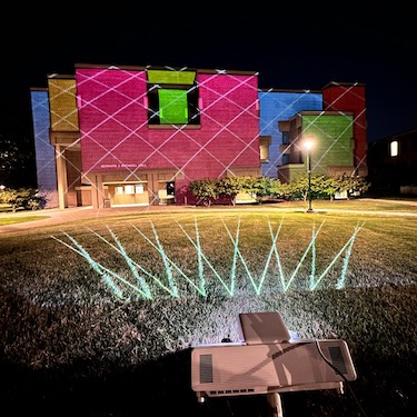 A photo of the exterior of Bernard J. Brommel Hall with a projector lighting the building with a rainbow of color blocks.