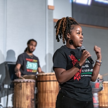 A person speaking at a past Northeastern Black History Month event, holding a microphone. Another person is visible in the background, playing drums. The speaker is wearing a black shirt with red, white and green graphics. The text on the shirt reads "Celebrating Black Resistance: Black History Month." 