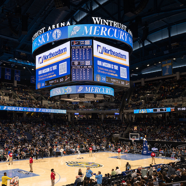 A photo of the jumbotron at Wintrust Arena during a Chicago Sky basketball game versus the Pheonix Mercury. The screens show the Northeastern Illinois University logo and tagline "Our Mission is You!" with a QR code to scan for more information. A crowd is visible in the stands in the background and players are visible on the court..