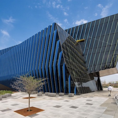 A photo of the exterior of NEIU's El Centro Location showing the blue and yellow louvers on a sunny day with a few clouds in the sky. A small tree is visible in the foreground and a person is walking near the building's entrance.