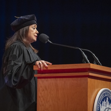 Iris Y. Martinez, wearing a black graduation cap and gown, delivering her speech at a podium with a microphone at Northeastern's December 2024 Commencement ceremony. 