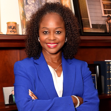 A photo of Dr. Katrina E. Bell-Jordan, smiling, wearing a bright blue blazer and white blouse with arms folded across her chest. A mantel with photos is visible in the background. 