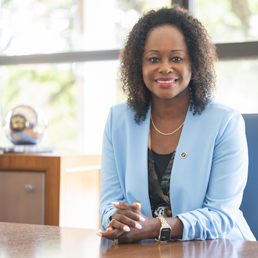 A photo of Katrina E. Bell-Jordan, Ph.D., wearing light blue blazer and dark colored blouse, smiling with hands folded on a wooden table. The NEIU Presidential Orb is visible in the background, with sunlight shining through windows. 