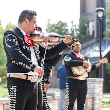 A photo of three musicians in a mariachi band wearing black suits with silver and gold embellishments, red ties and white collared shirts, perform on Northeastern's University Commons on a sunny day. The Ronald Williams Library is visible in the background. 