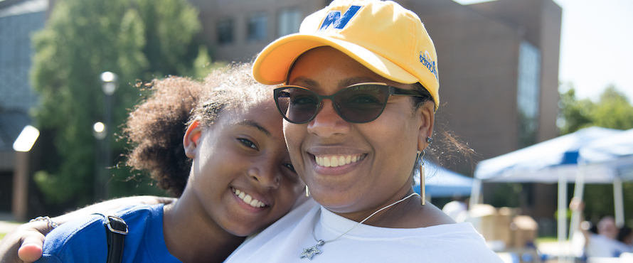 A person in a blue NEIU shirt and a person in a white N logo shirt and yellow N logo hat in a warm embrace smiling in a photo at a NEIU Weekend 2019