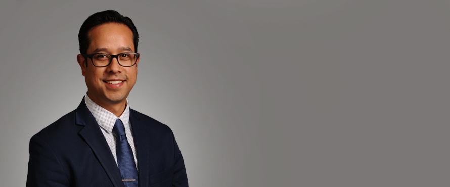 A headshot photo of Aaron Cortes wearing dark colored suit with blue tie and collard shirt and glasses, smiling against a gray background.