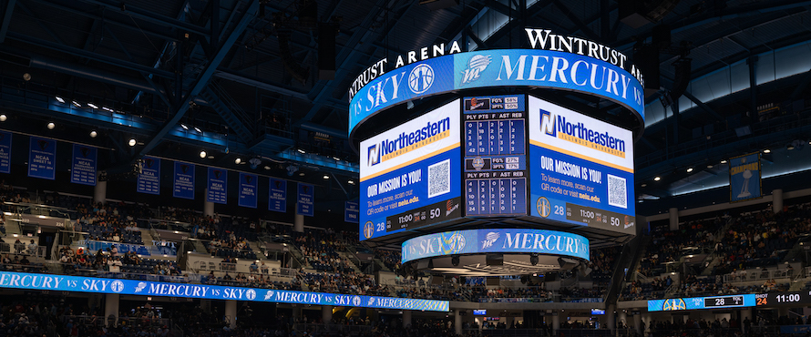 A photo of the jumbotron at Wintrust Arena during a Chicago Sky basketball game versus the Pheonix Mercury. The screens show the Northeastern Illinois University logo and tagline "Our Mission is You!" with a QR code to scan for more information. A crowd is visible in the stands in the background.