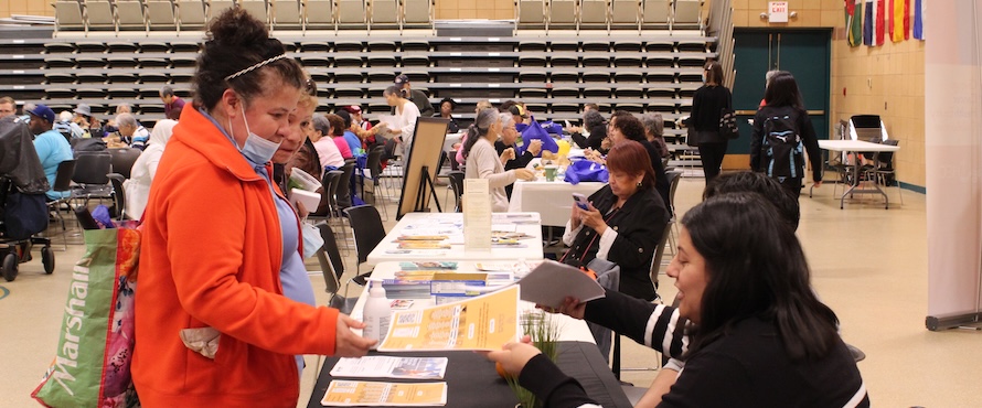 A person in an orange coat is handed a flyer from a person in a black and white striped sweater sitting behind a table during the ChicagoCHEC Community Forum in 2023. 