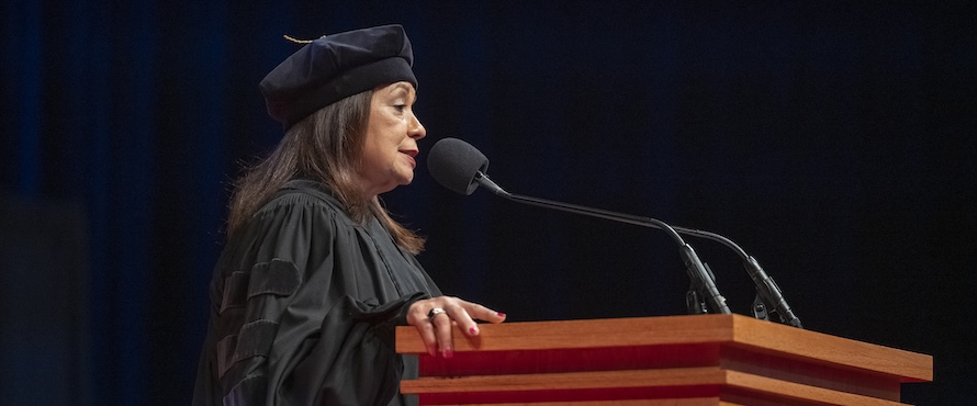 Iris Y. Martinez, wearing a black graduation cap and gown, delivering her speech at a podium with a microphone at Northeastern's December 2024 Commencement ceremony. 