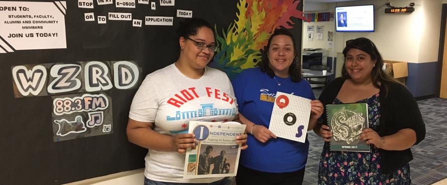 Students from the Independent, Que Ondee Sola and Seeds pose with their publications in front of the WZRD radio bulletin board.